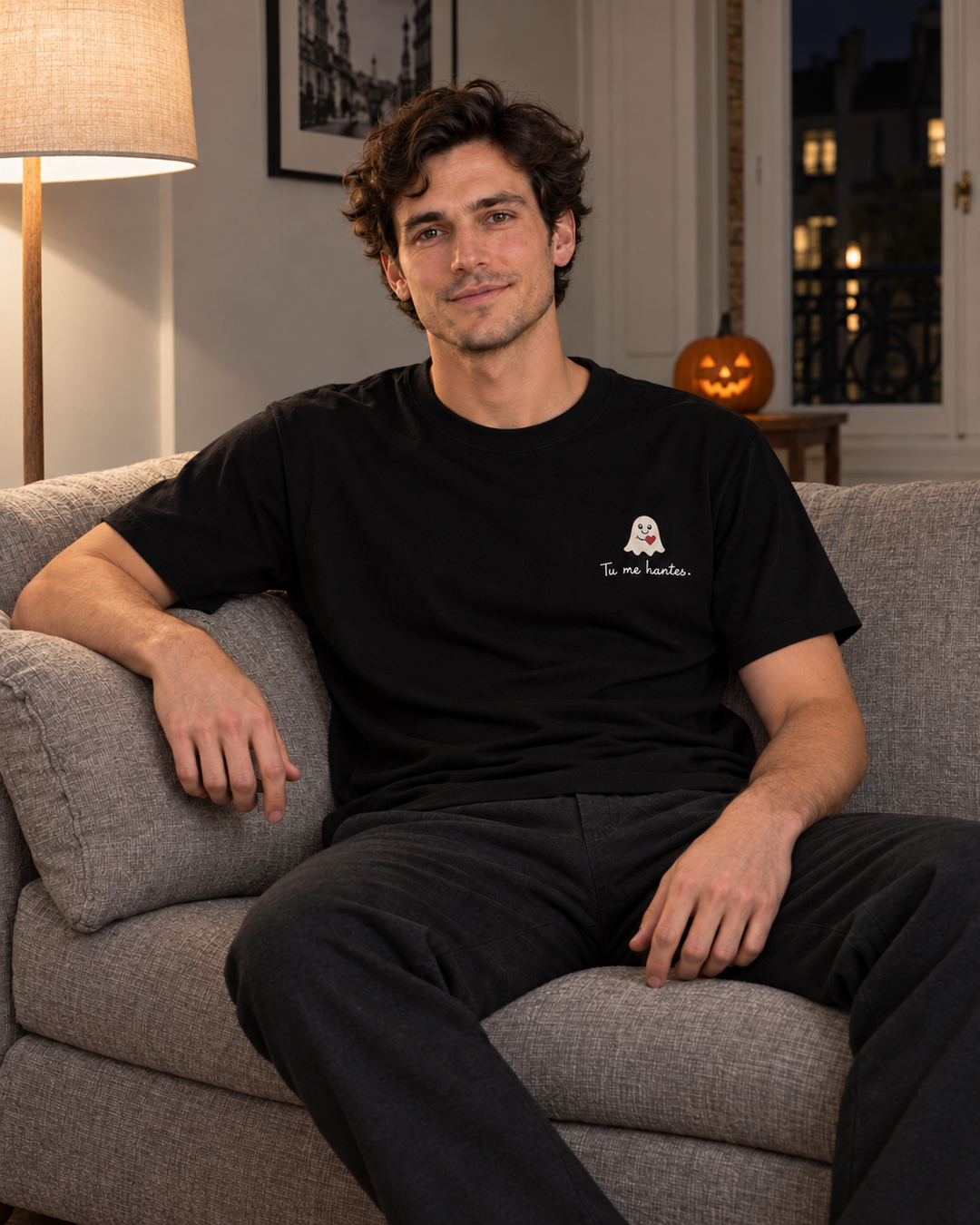 Simulation IA du t-shirt noir brodé « Tu me hantes. », porté par un homme assis dans un canapé, décor Halloween de nuit — Rufus Paris.