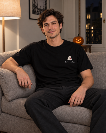 Simulation IA du t-shirt noir brodé « Tu me hantes. », porté par un homme assis dans un canapé, décor Halloween de nuit — Rufus Paris.